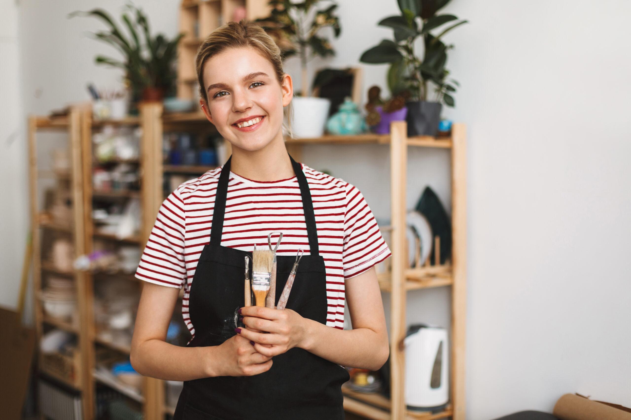 cheerful-girl-black-apron-striped-t-shirt-holding-pottery-tools-hands-happily-looking-camera-pottery-studio